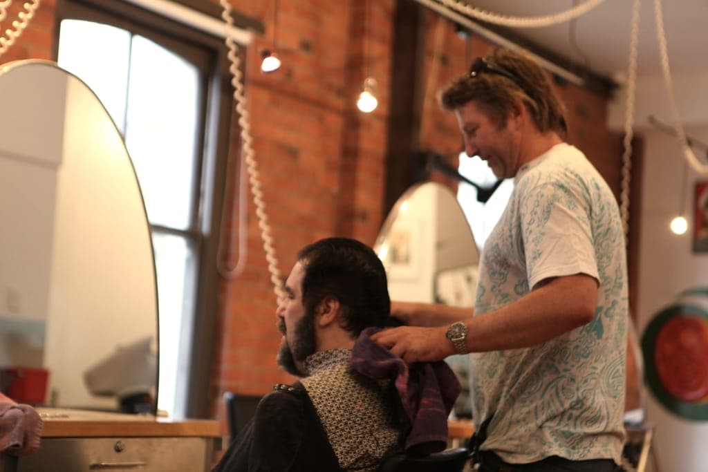Barbier qui rit en finissant la coupe d'un client barbu, mur de briques et grande fenêtre