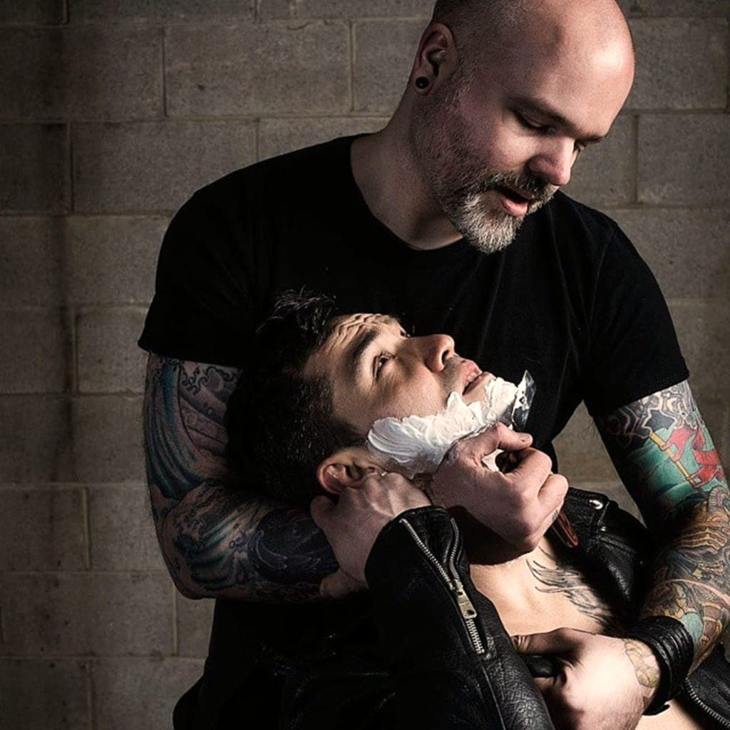 Barbier tatoué qui rase un client au coupe-chou, mousse sur la joue, tête renversée