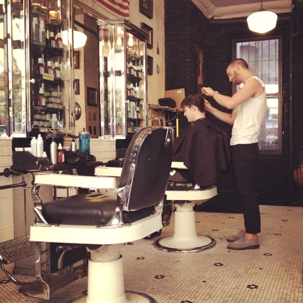 Barbier au travail derrière un client, fauteuils chromés et étagères de flacons