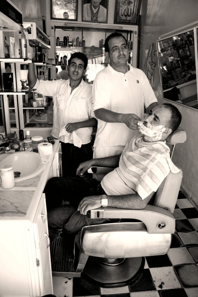 Photo noir et blanc : client renversé dans le fauteuil, visage couvert de mousse, barbier penché sur lui